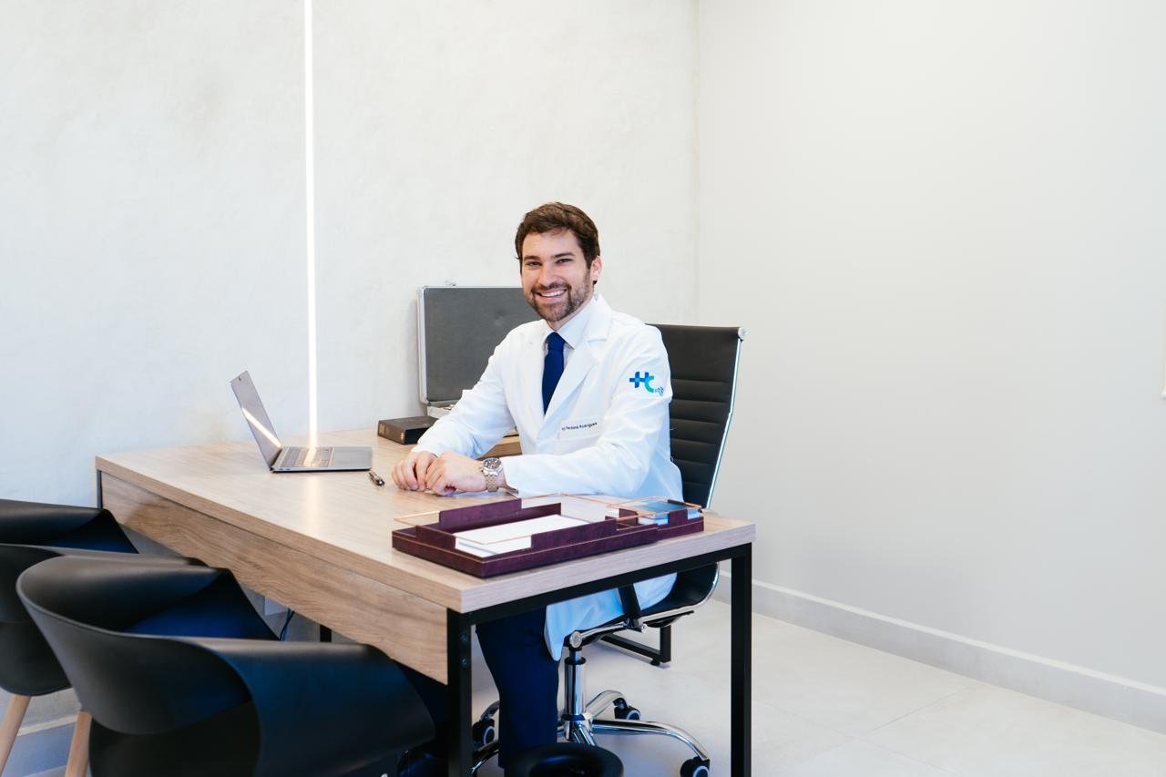 Dr. Iago sentado à mesa do consultório, sorrindo.